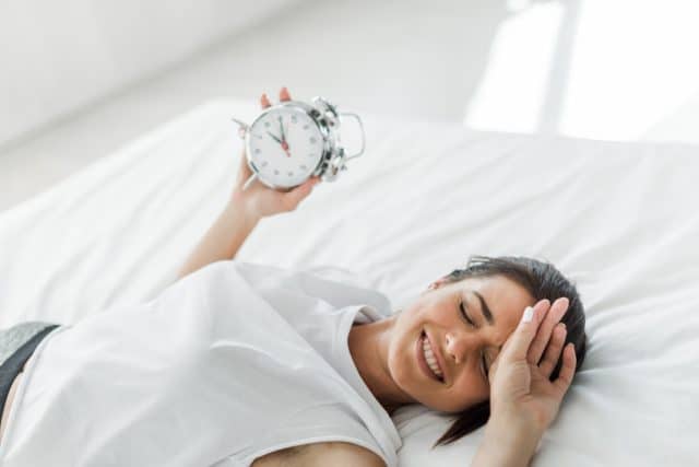 Happy woman holding an alarm clock in bed in the morning